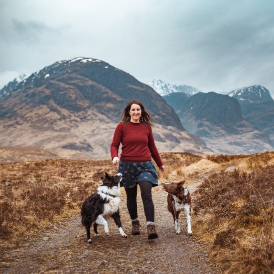 A photo of a woman with her two border collies with the mountain scenery of Glencoe behind them