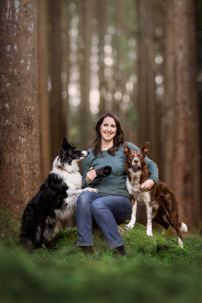 A photographer in the woods with her two border collies, holding a camera, fine art photography style
